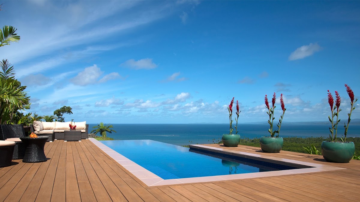 Infinity pool overlooking blue ocean and sky at Emaho Sekawa Retreat Fiji, with wooden deck, potted red flowers, and lounge seating.