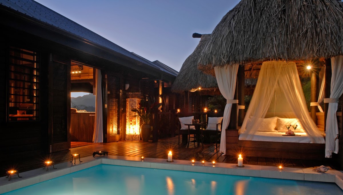 Emaho Sekawa Retreat Fiji villa at dusk with illuminated pool, thatched-roof bungalow, wooden deck, candlelit lounge area, and mountain views beyond.