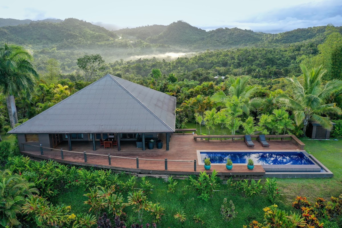 Modern pavilion with peaked gray roof and wooden deck overlooking a pool, surrounded by tropical gardens and forested mountains in Fiji.