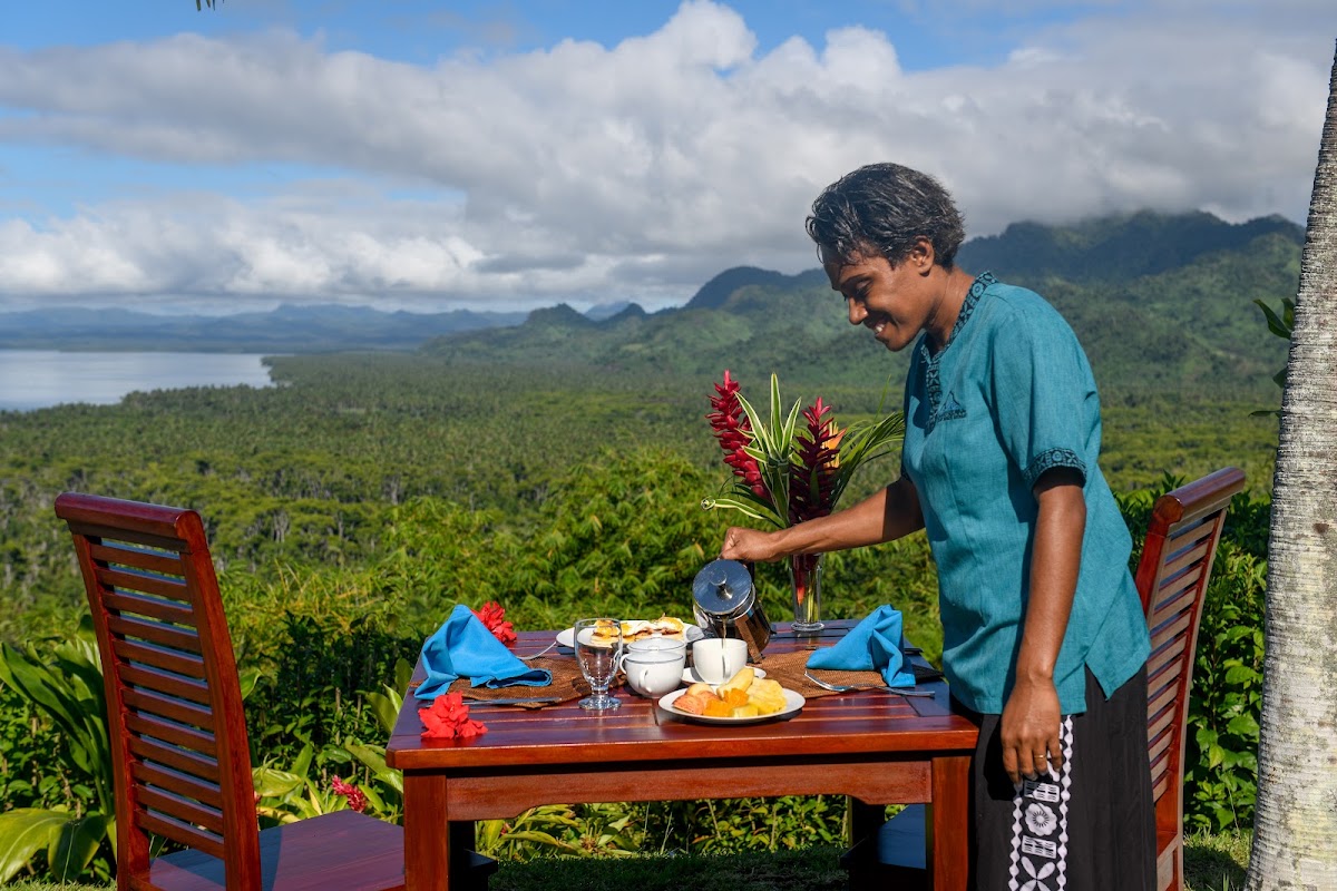 Server arranges breakfast at a clifftop table overlooking rainforest, mountains, and distant lake under cloudy sky.