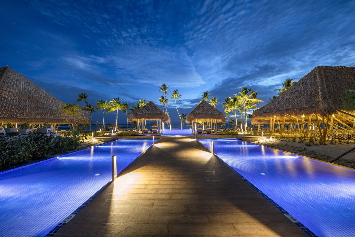 Lit walkway between glowing blue pools leading to thatched-roof bungalows and beach at dusk.