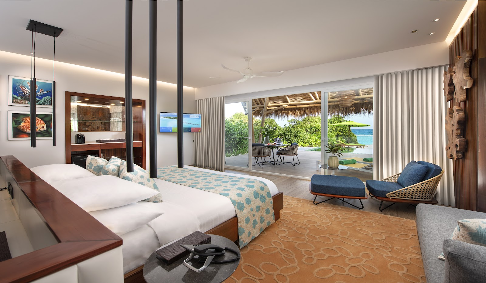 Resort suite with ocean view through glass doors, featuring king bed, blue lounge chair, and direct access to beachfront patio with thatched-roof structures visible beyond.