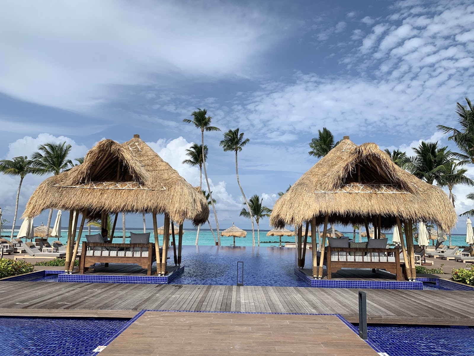 Two thatched-roof cabanas with lounge seating overlook a lagoon pool at Emerald Maldives Resort & Spa, with palm trees and turquoise ocean beyond.