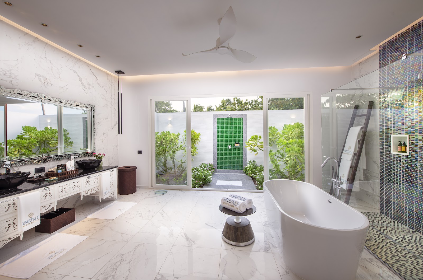 Spa bathroom with freestanding white tub, marble countertops, green tile shower, and glass doors opening to a private courtyard with tropical plants.