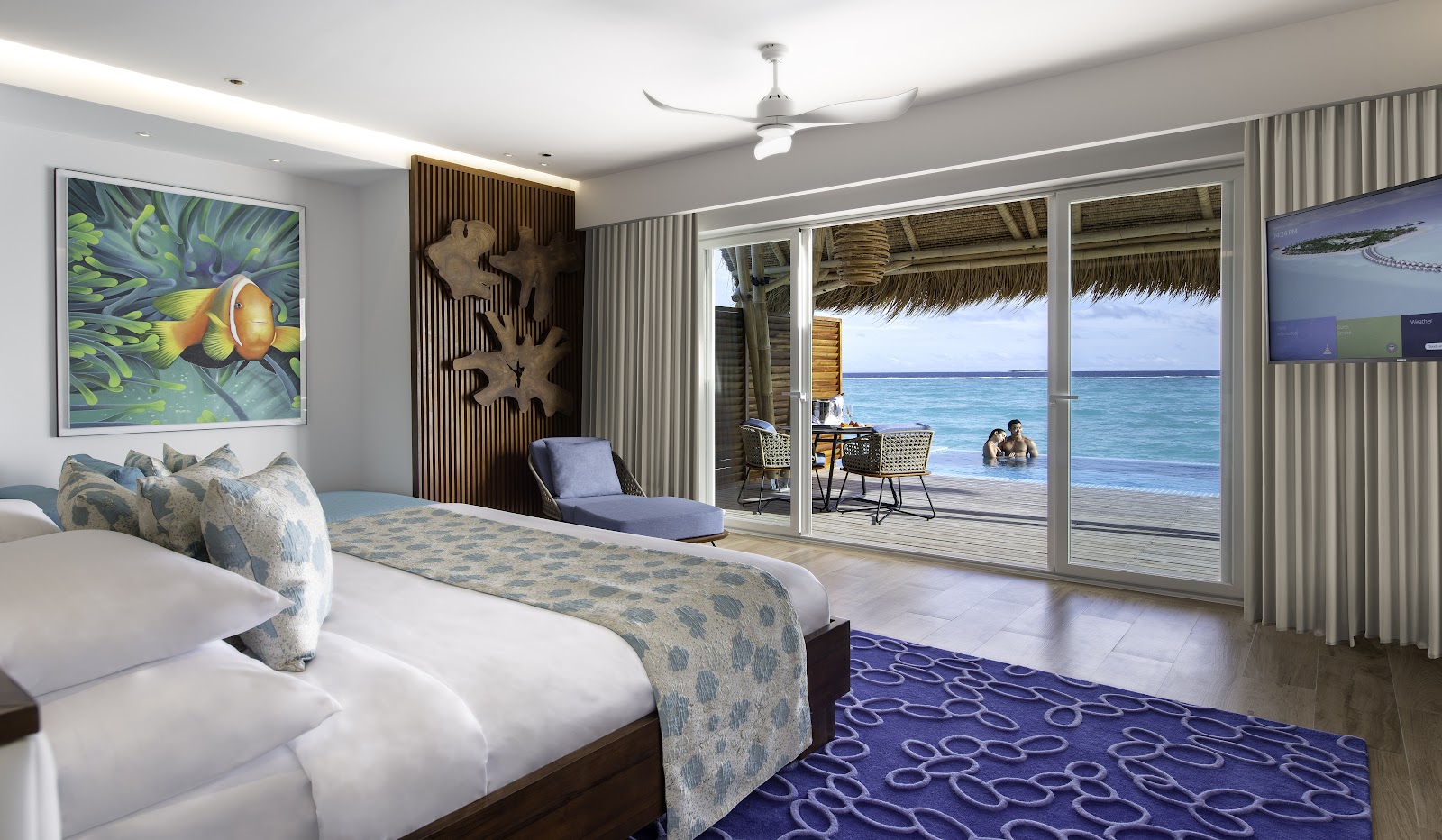 Beachfront suite at Emerald Maldives Resort & Spa with ocean view through glass doors, bed with patterned runner, and guests wading in turquoise water beyond the deck.