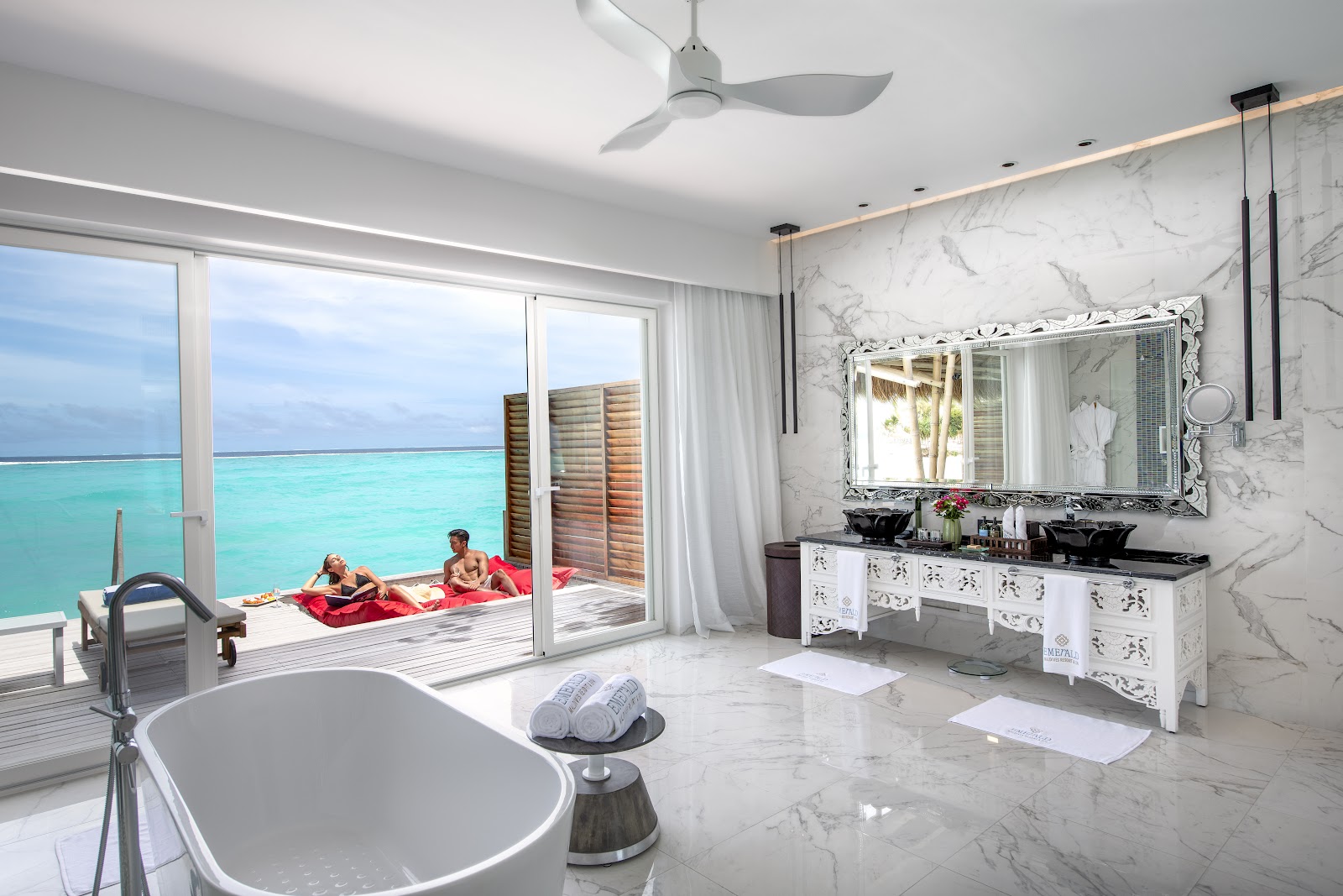 Luxury bathroom with marble walls and freestanding tub overlooking ocean-view deck where guests relax on red loungers at Emerald Maldives Resort & Spa.