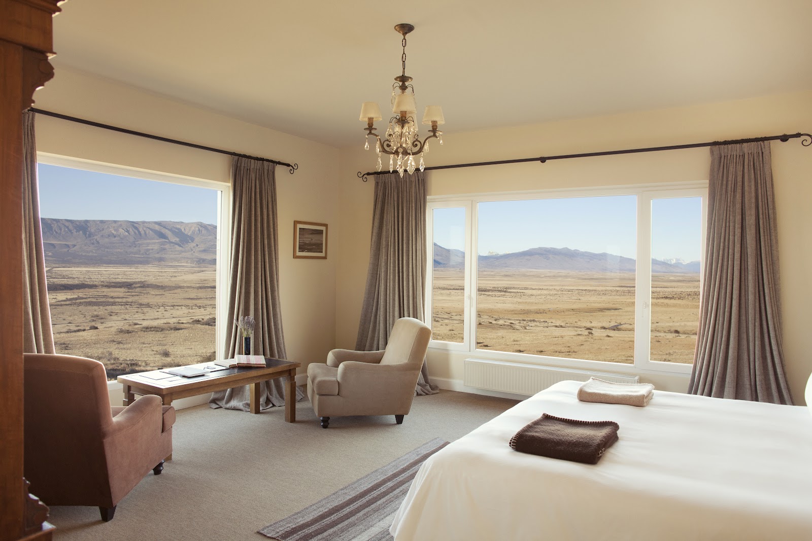 Spacious resort bedroom with cream walls, a bed with brown throw, upholstered chairs, and large windows overlooking flat desert landscape and distant mountains.