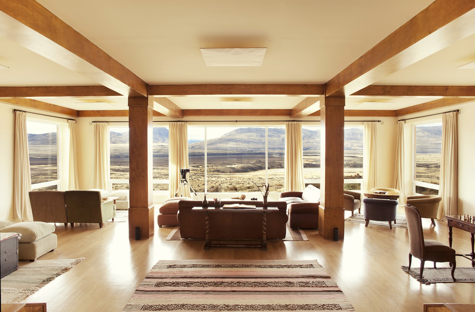 Spacious lounge with brown leather seating, wooden beams, and floor-to-ceiling windows framing expansive Patagonian valley and mountain views at EOLO Patagonia's Spirit.