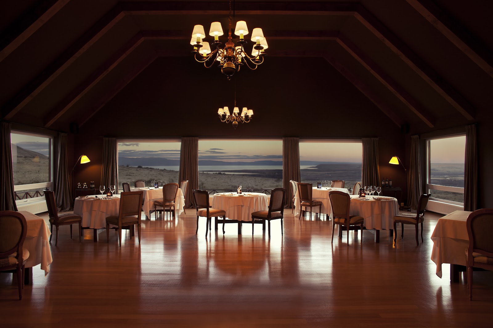 Fine dining room at EOLO Patagonia's Spirit with round tables, panoramic windows overlooking vast landscape at dusk, and brass chandeliers.