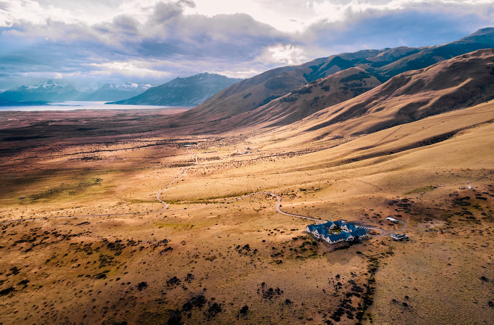 A blue lodge sits isolated on golden grassland below snow-capped mountains and a turquoise lake in Argentine Patagonia.