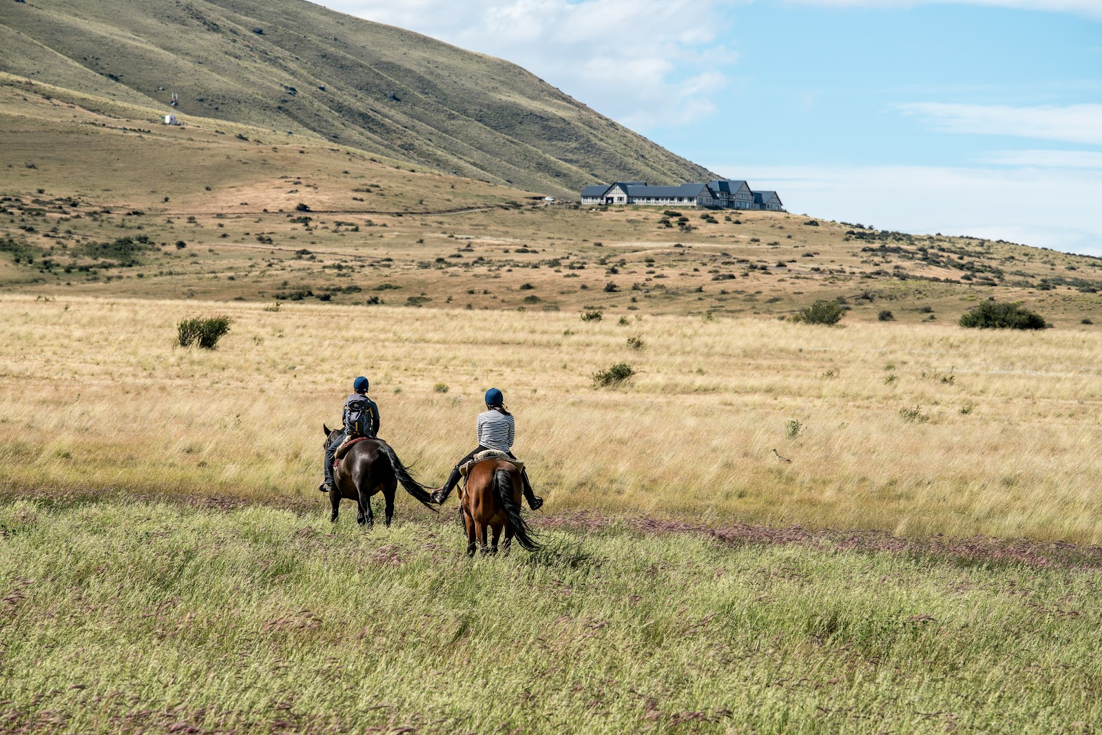 Two riders on horseback in grassland with a dark-roofed lodge nestled against a steep hillside in Patagonia.