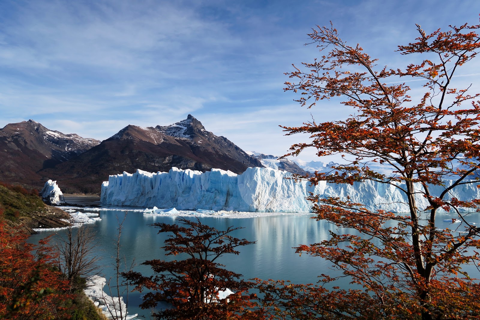 Glacier with blue-white ice wall reflected in calm lake water, framed by reddish autumn trees and dark mountains under blue sky.