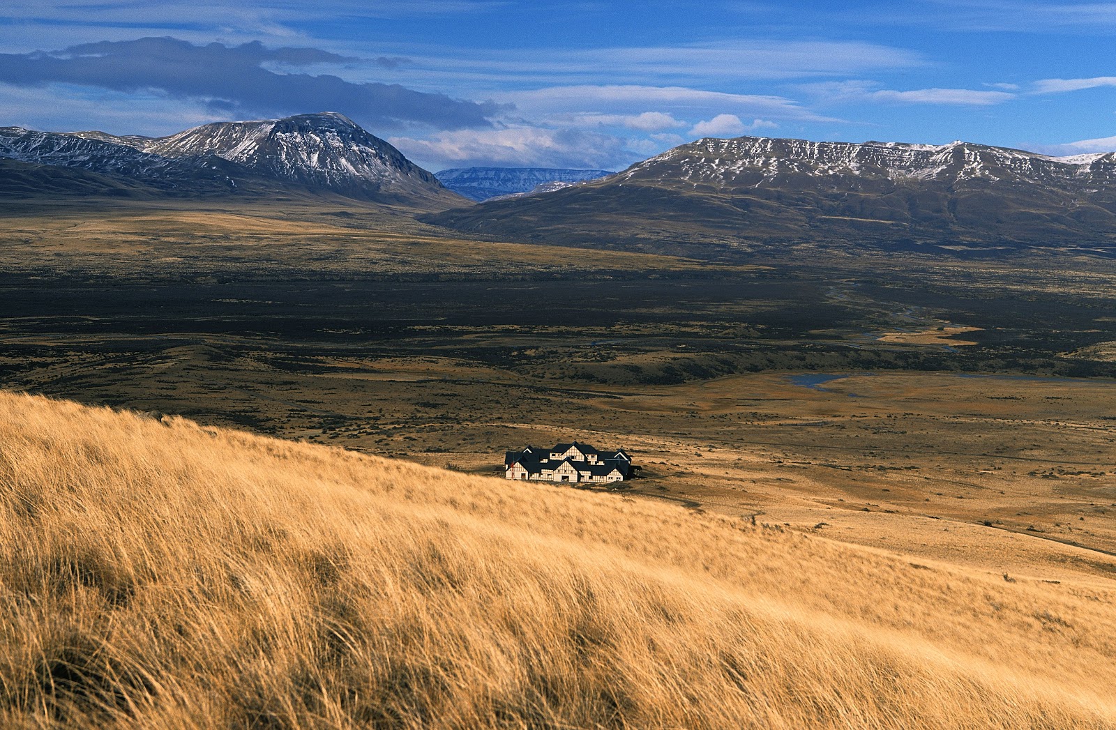 EOLO Patagonia's Spirit sits in a vast valley surrounded by snow-capped mountains under blue sky with golden grassland in foreground.