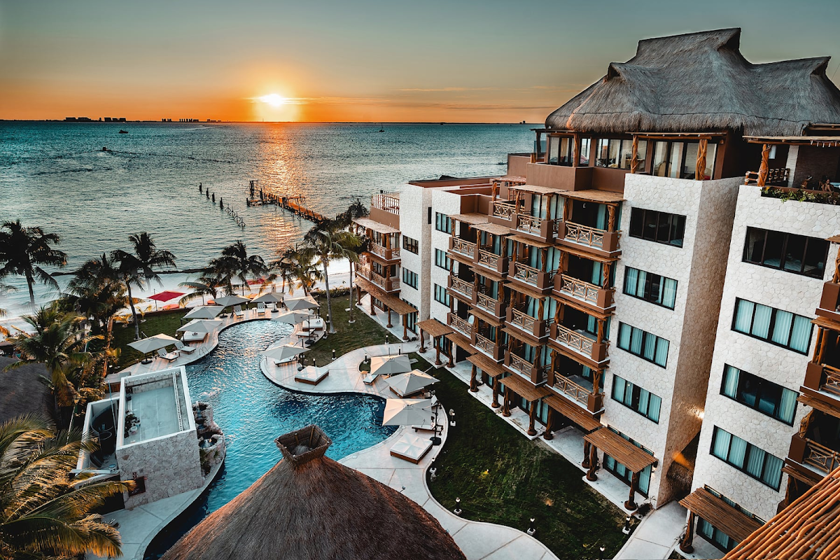 Beachfront resort buildings with curved pool overlooking turquoise water and sunset on the horizon.