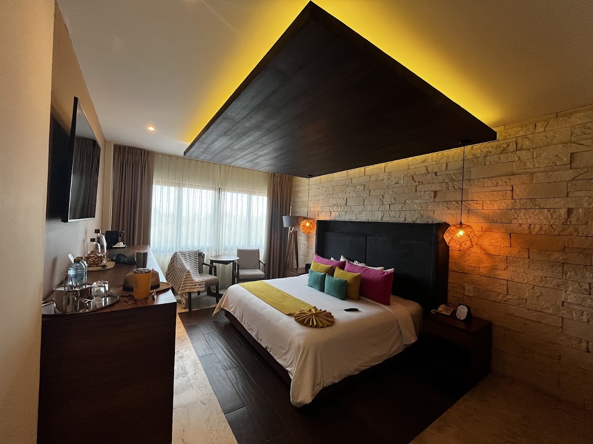Modern resort bedroom with stone accent wall, black canopy bed with colorful pillows, amber pendant lights, and seating area with large windows.