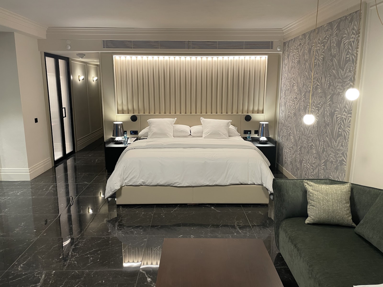 Modern resort bedroom with king bed, white linens, black marble floors, illuminated headboard, patterned wallpaper, and seating area with green sofa.