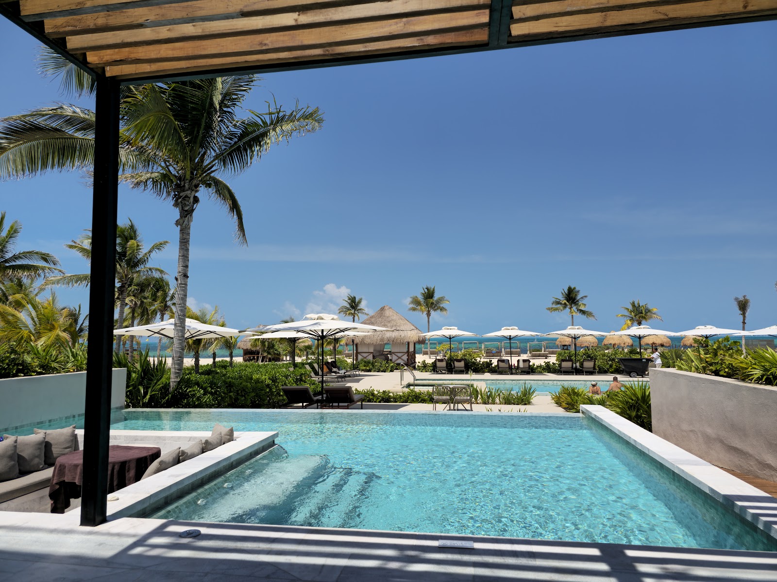 Resort pool with wooden pergola shade overlooking beach umbrellas, palms, and ocean under clear blue sky.