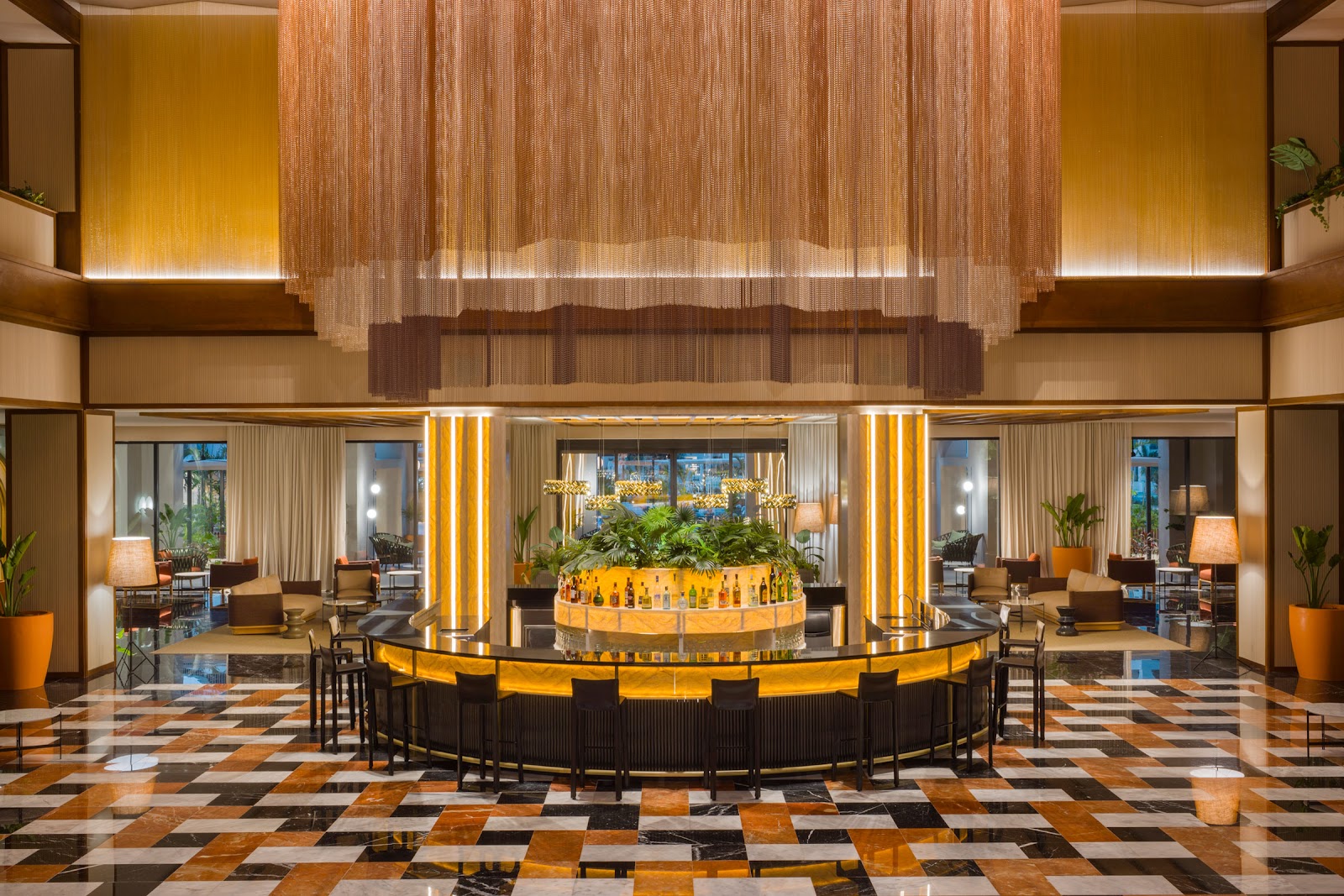 Resort lobby bar with circular yellow and black counter, geometric floor, hanging copper curtains, and potted plants under warm lighting.