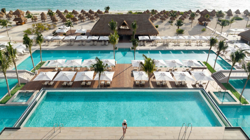 Aerial view of Excellence Coral Playa Mujeres with multiple pools, loungers, thatched bungalows, and turquoise Caribbean waters beyond.
