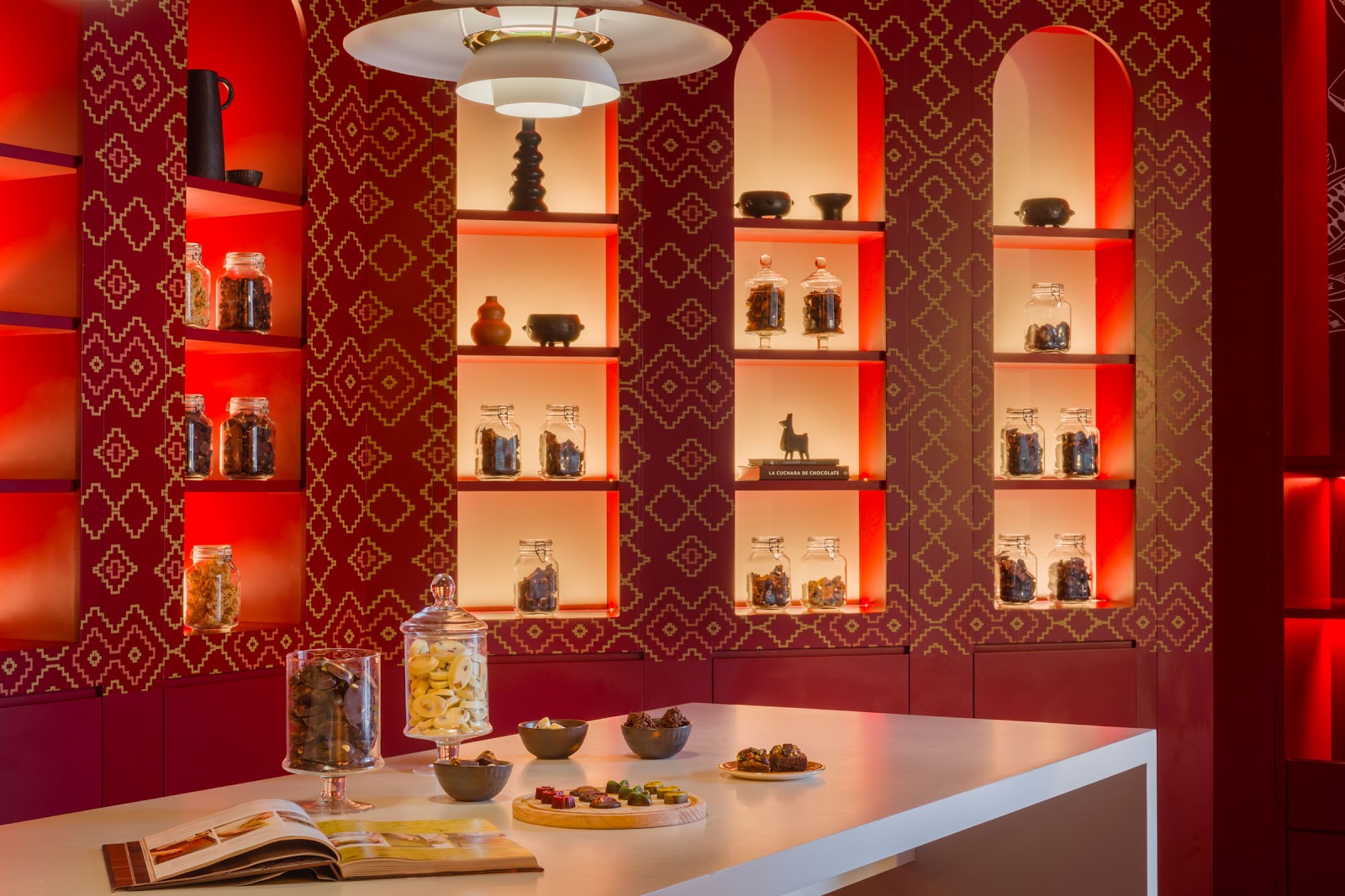 Modern resort spa or wellness space with red and gold geometric wallpaper, illuminated shelving displaying glass jars, and a white counter with food and beverage service.