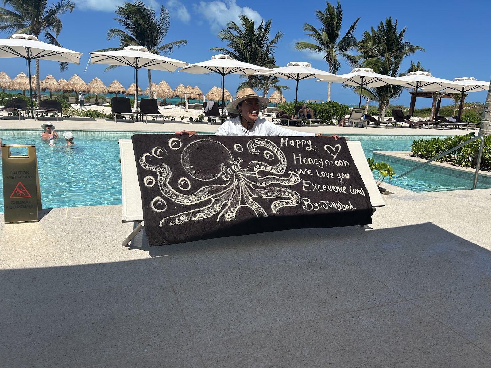 Staff member holds a chalkboard sign with octopus drawing at Excellence Coral Playa Mujeres poolside, with palm trees and thatched cabanas beyond.