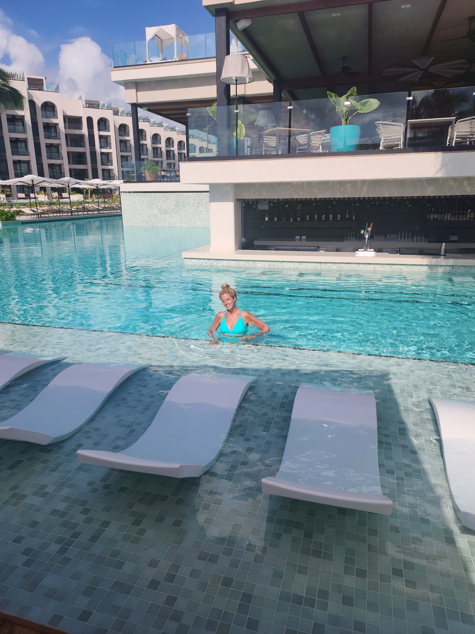 Woman in turquoise swimsuit wades in pool at Excellence Coral Playa Mujeres, with white loungers in foreground and modern resort buildings beyond.