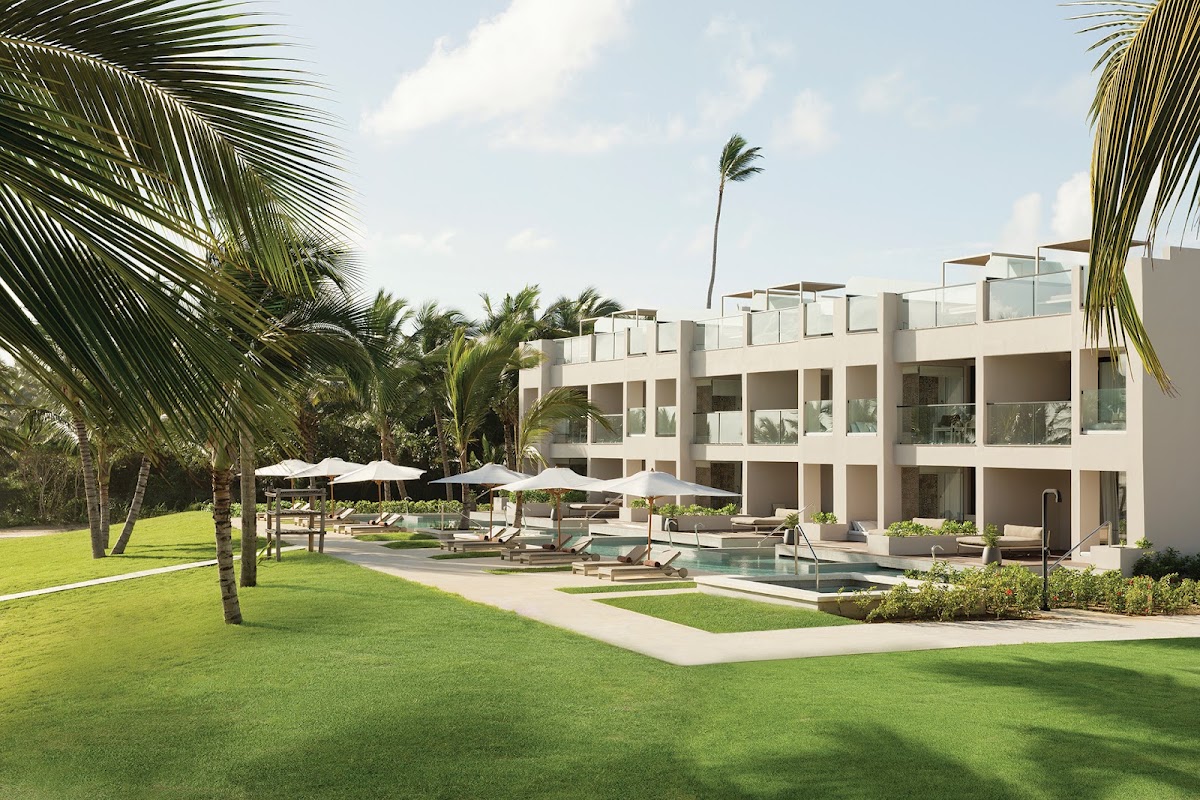 White multi-story resort building with pool, loungers, umbrellas, and palm trees on manicured lawn at Excellence El Carmen.