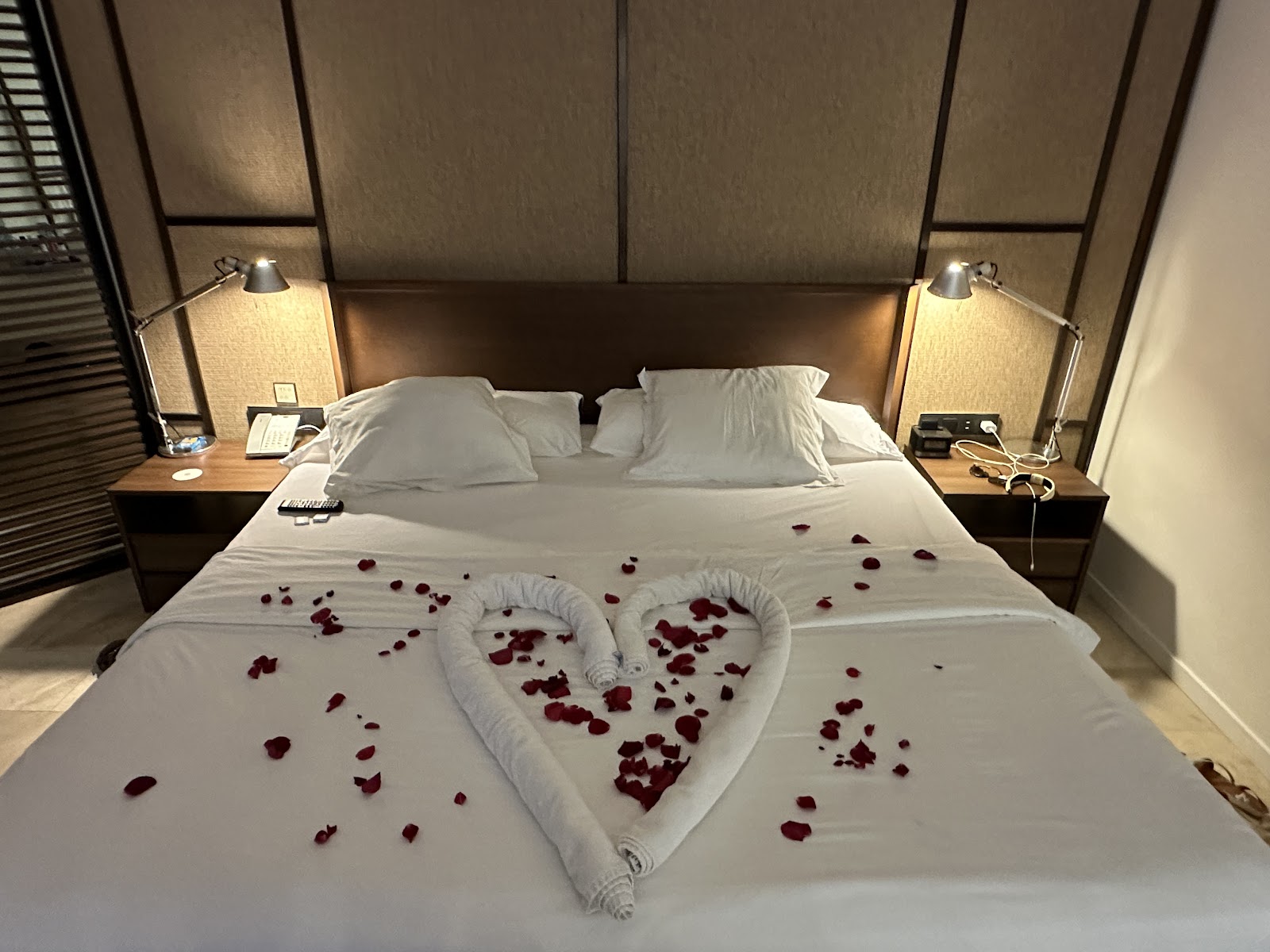 A bed at Excellence El Carmen decorated with a heart-shaped towel and red rose petals, flanked by bedside tables with lamps.