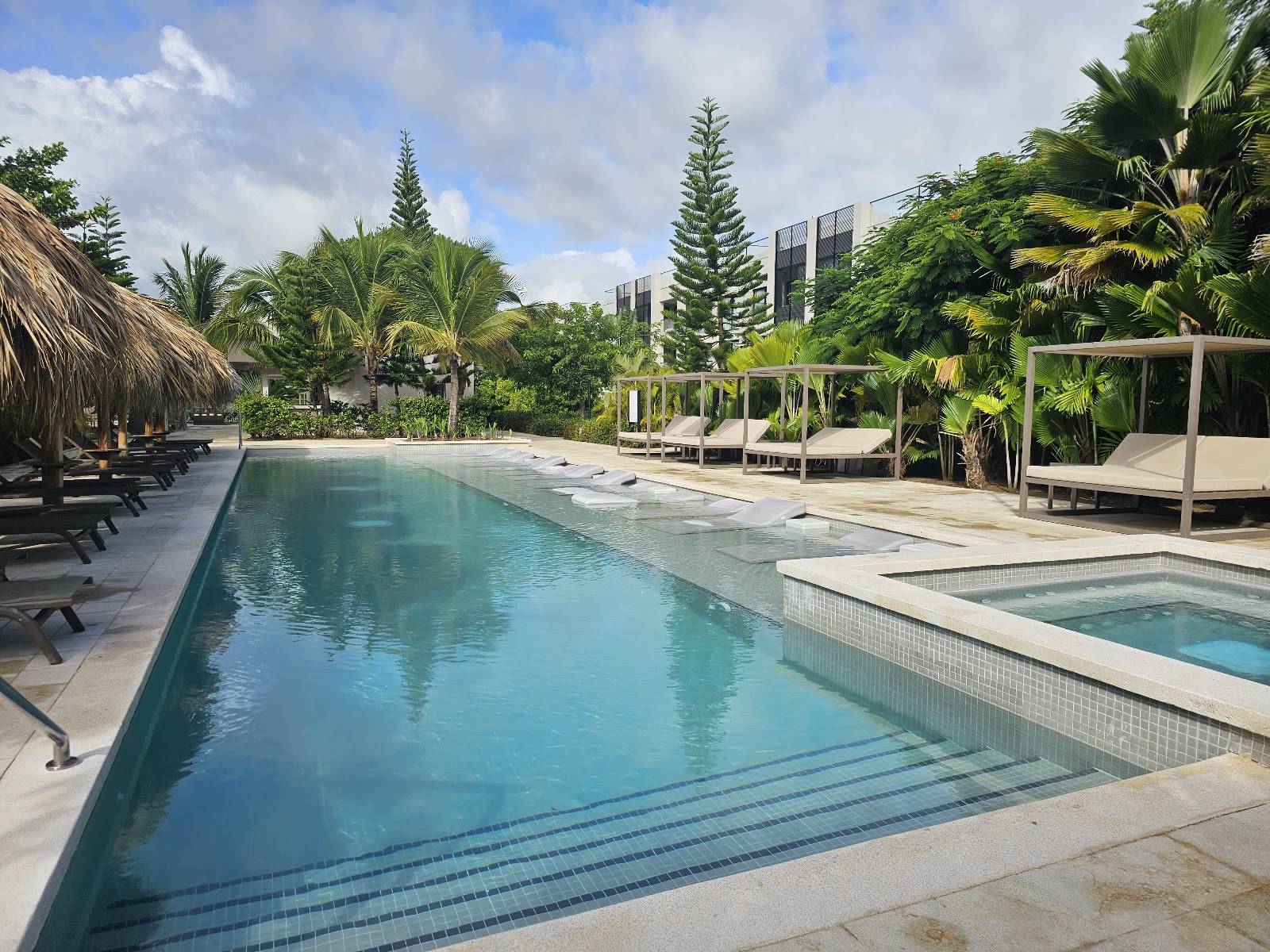 Long lap pool with lane markings beside a hot tub at Excellence El Carmen, surrounded by lounge chairs, thatched palapas, and tropical palms.