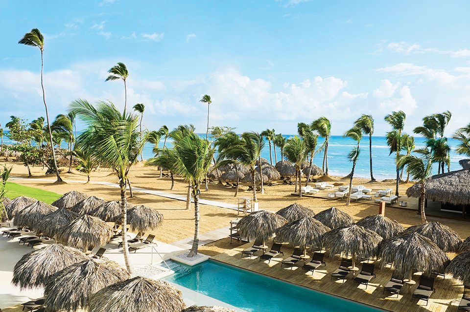 Resort pool with thatched umbrellas and lounge chairs overlooks palm-lined beach and turquoise ocean under blue sky.