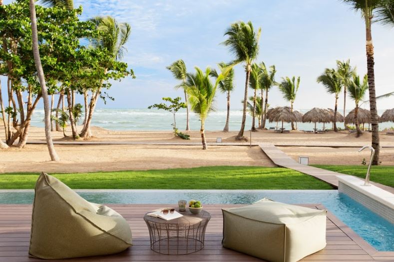 Bean bag chairs on a wooden deck with a pool, manicured lawn, palm trees, and beach with thatched umbrellas beyond.