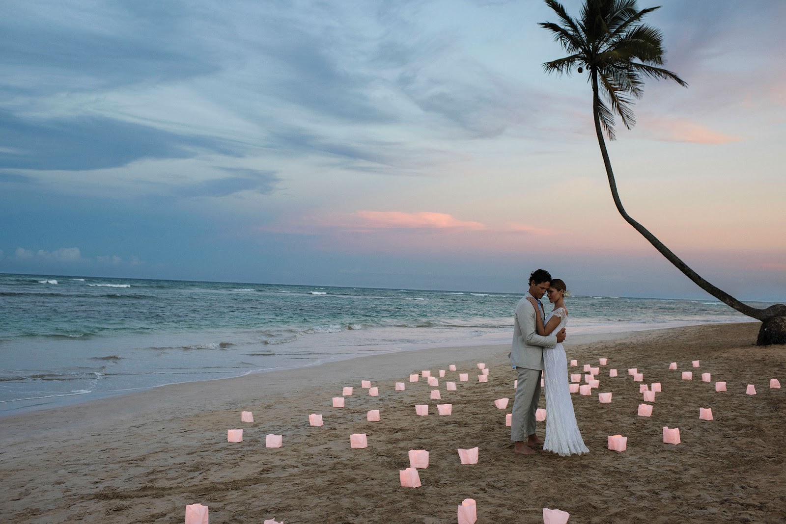 Couple in wedding attire embrace on a beach at sunset surrounded by lit pink candles, with a palm tree and ocean waves visible.