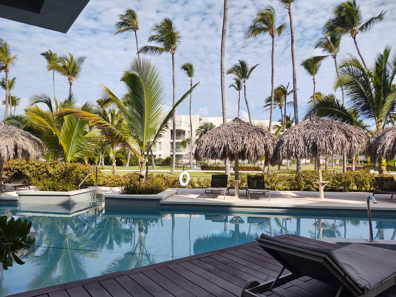 Resort pool with thatched palapas, palm trees, lounge chairs, and modern white building beyond under blue sky.