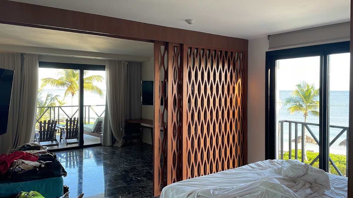 Resort bedroom with ocean view from balcony, wooden room divider, and palm trees visible through glass doors.
