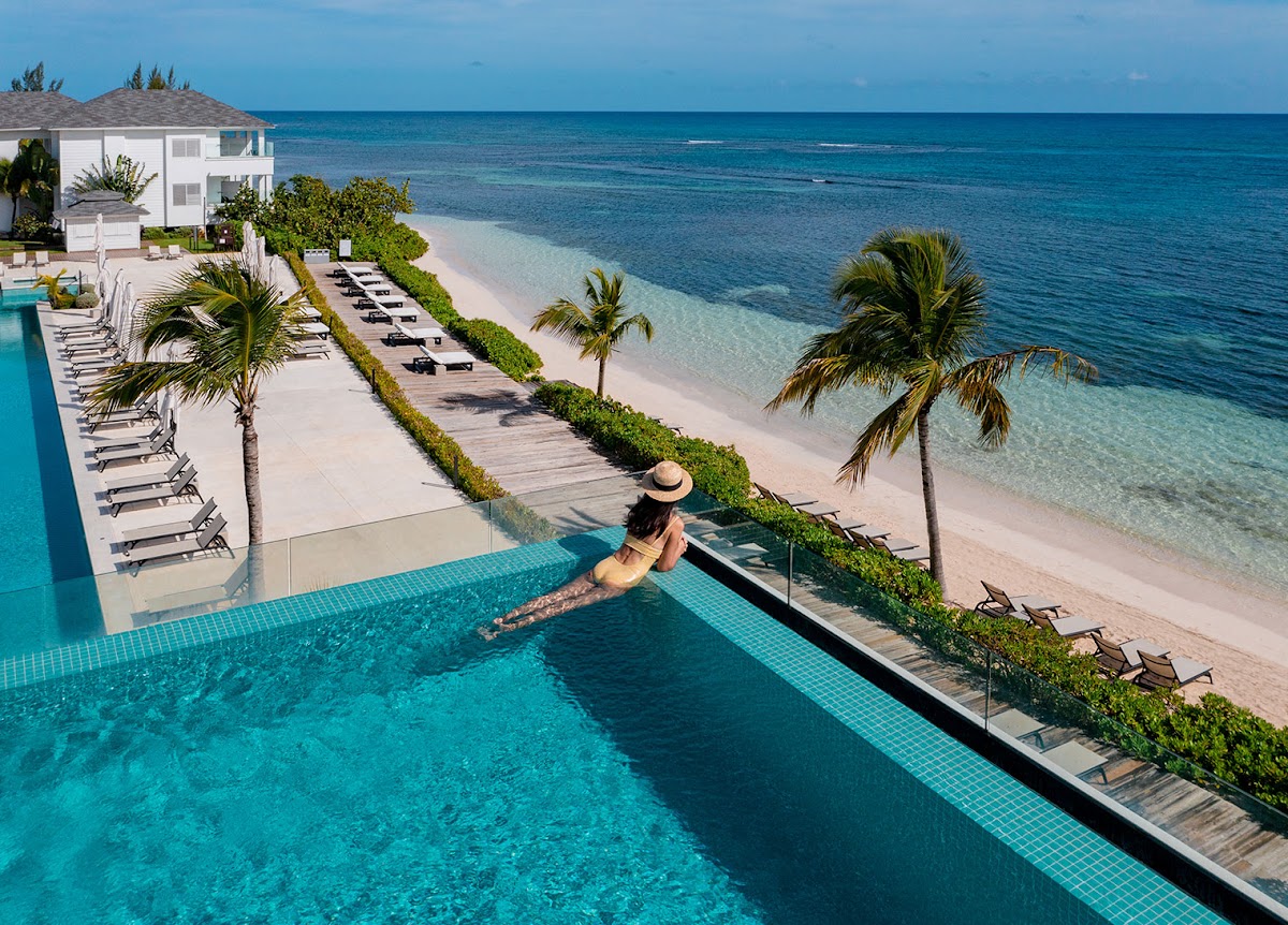 Guest swimming in infinity pool overlooking white sand beach, palm trees, and turquoise ocean at Excellence Oyster Bay.