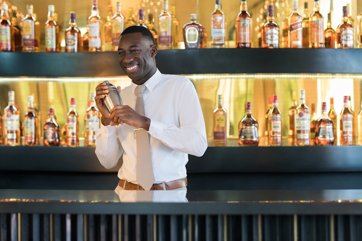 Bartender in white shirt holding a cocktail shaker behind a bar lined with amber and clear liquor bottles.