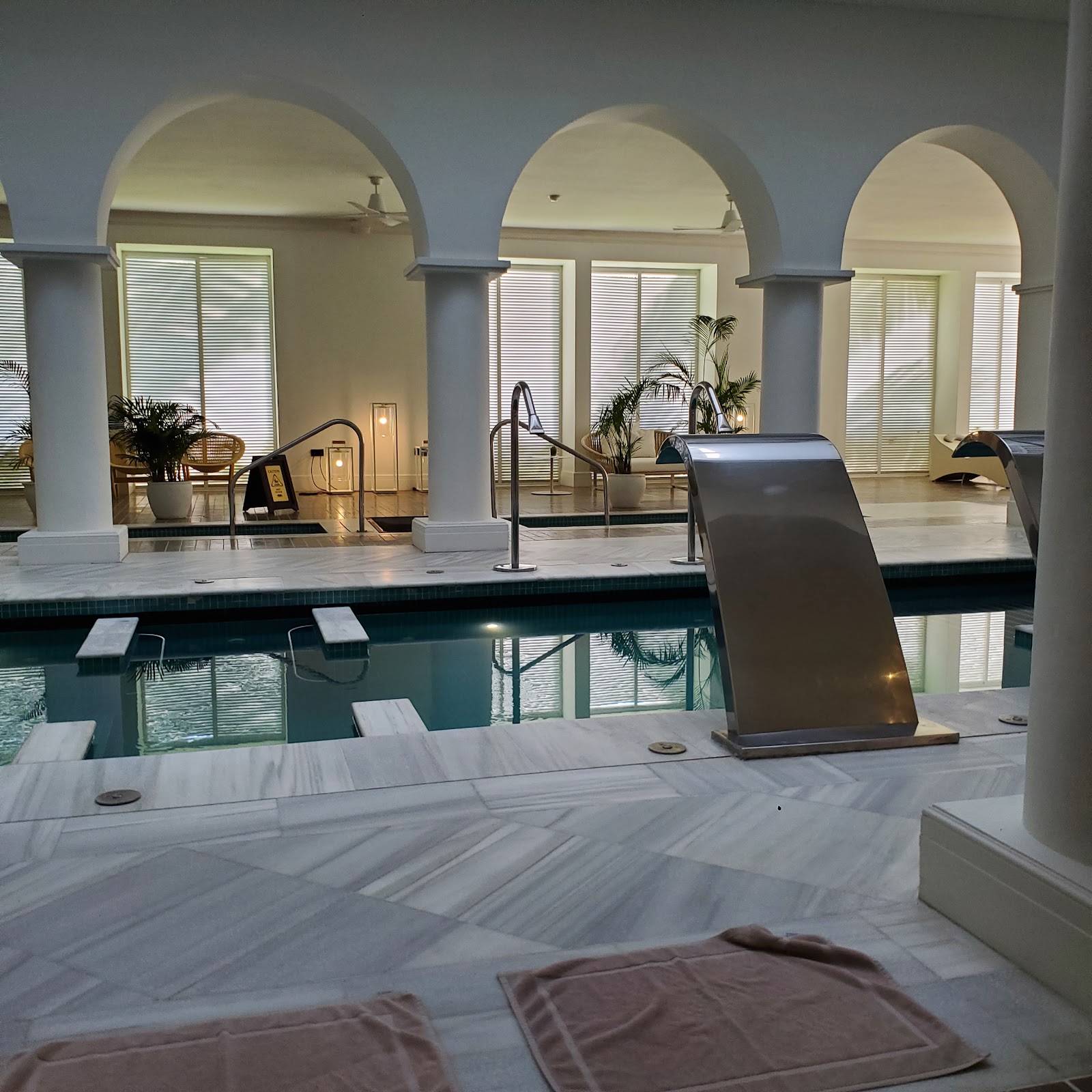 Indoor pool area at Excellence Oyster Bay with arched ceilings, white columns, lounge chairs, and water slides flanking the teal-colored pool.