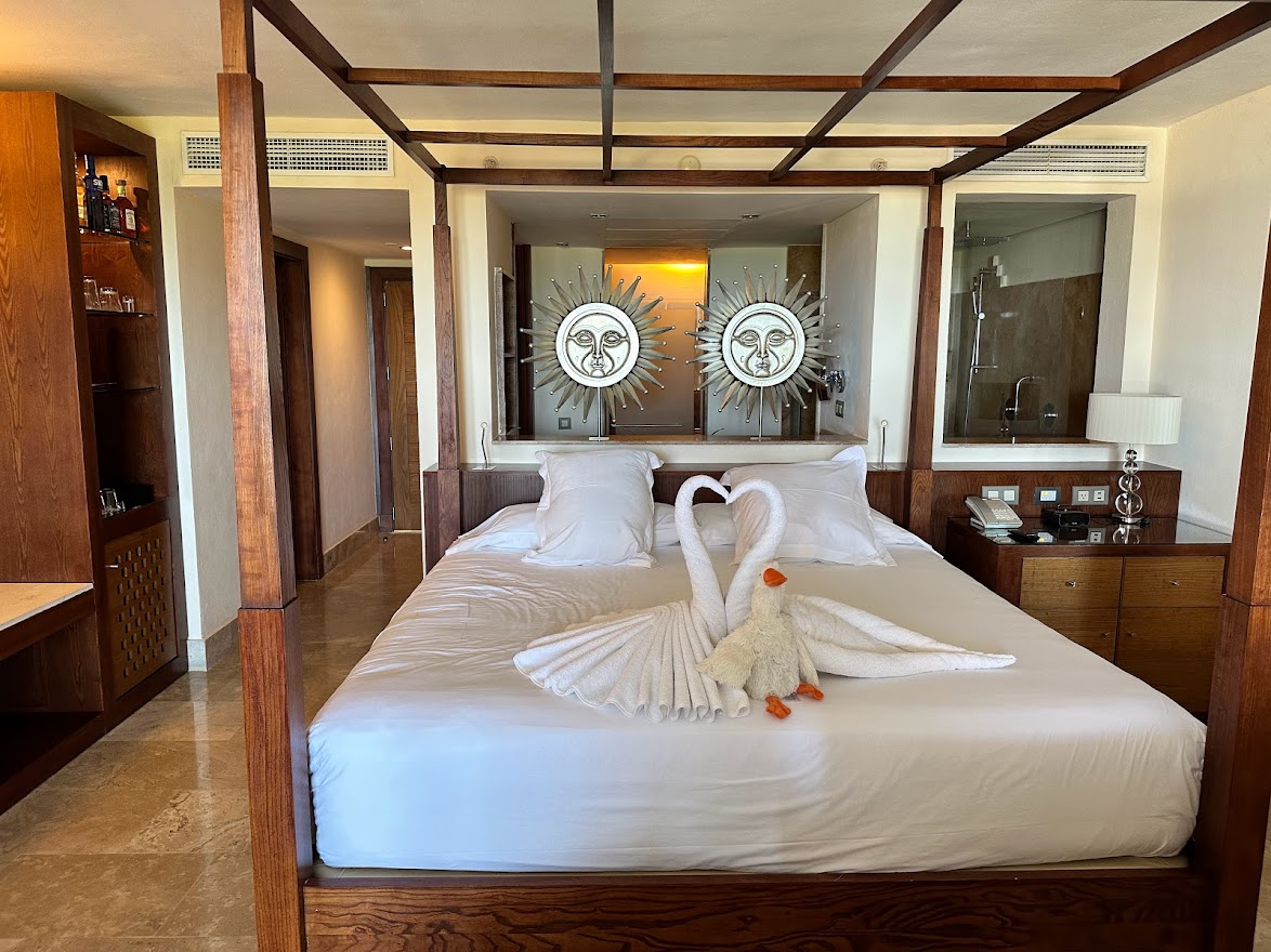 Four-poster bed with towel swan decoration in a spacious bedroom with wooden beams, sun-face wall art, and an ensuite bathroom at Excellence Playa Mujeres.