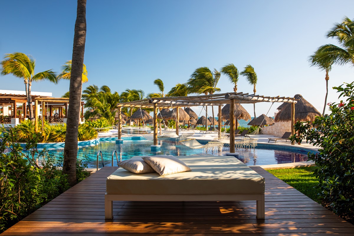 Wooden lounge bed with pillows on a deck overlooking the pool at Excellence Playa Mujeres, with palm trees and thatched palapas beyond.