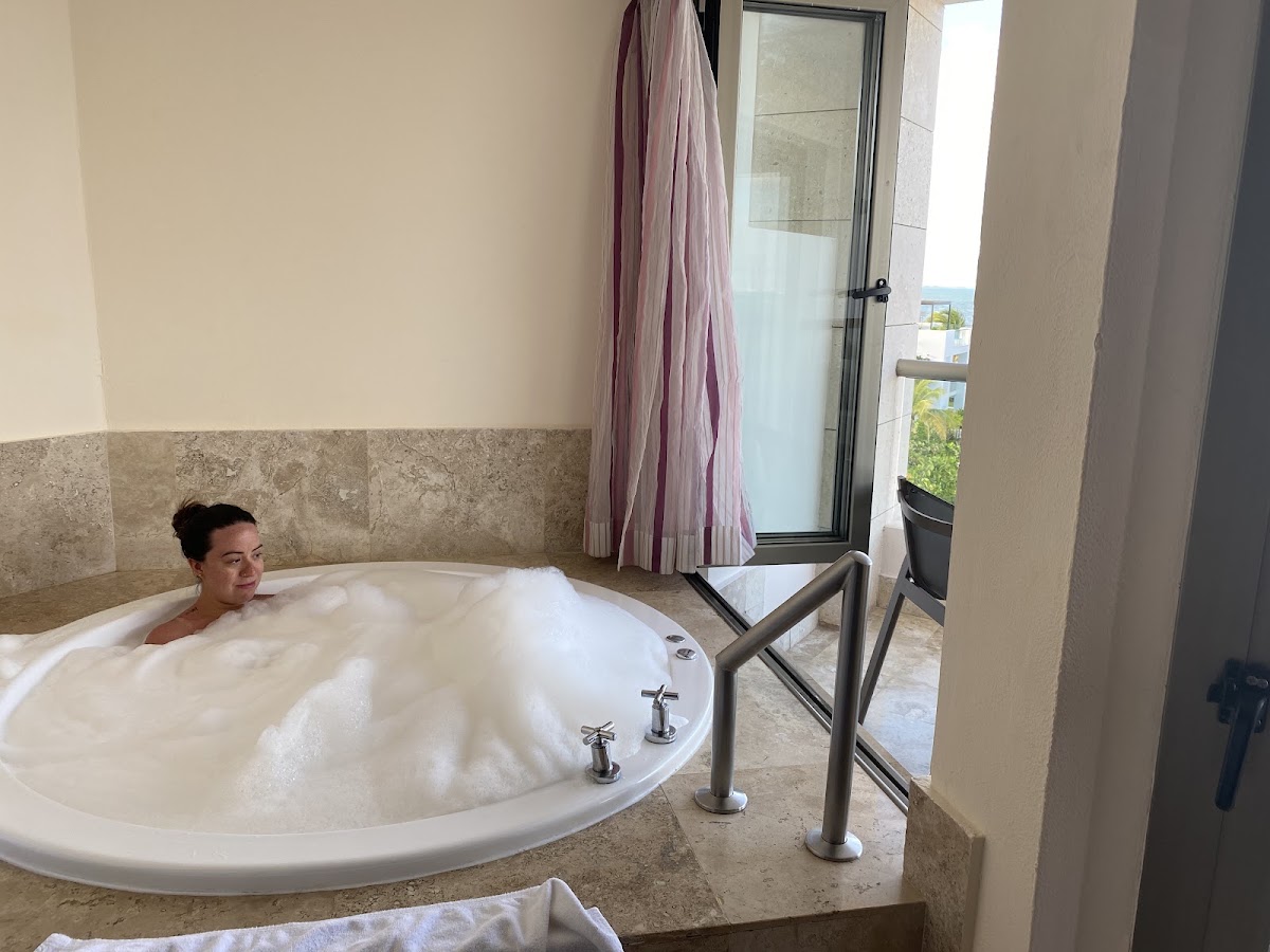 Woman relaxing in a large circular bathtub with jets at Excellence Playa Mujeres, with ocean view through the glass door.