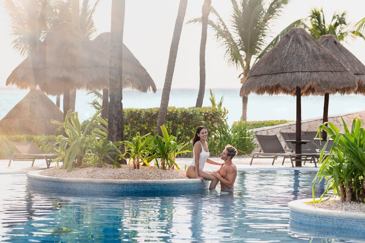Couple wading in a resort pool with thatched palapas, palm trees, and ocean view at Excellence Playa Mujeres.