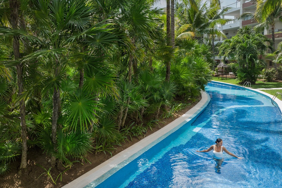 A guest swims in a curved blue pool flanked by lush palm trees and tropical plants at Excellence Playa Mujeres, with resort buildings visible in the background.