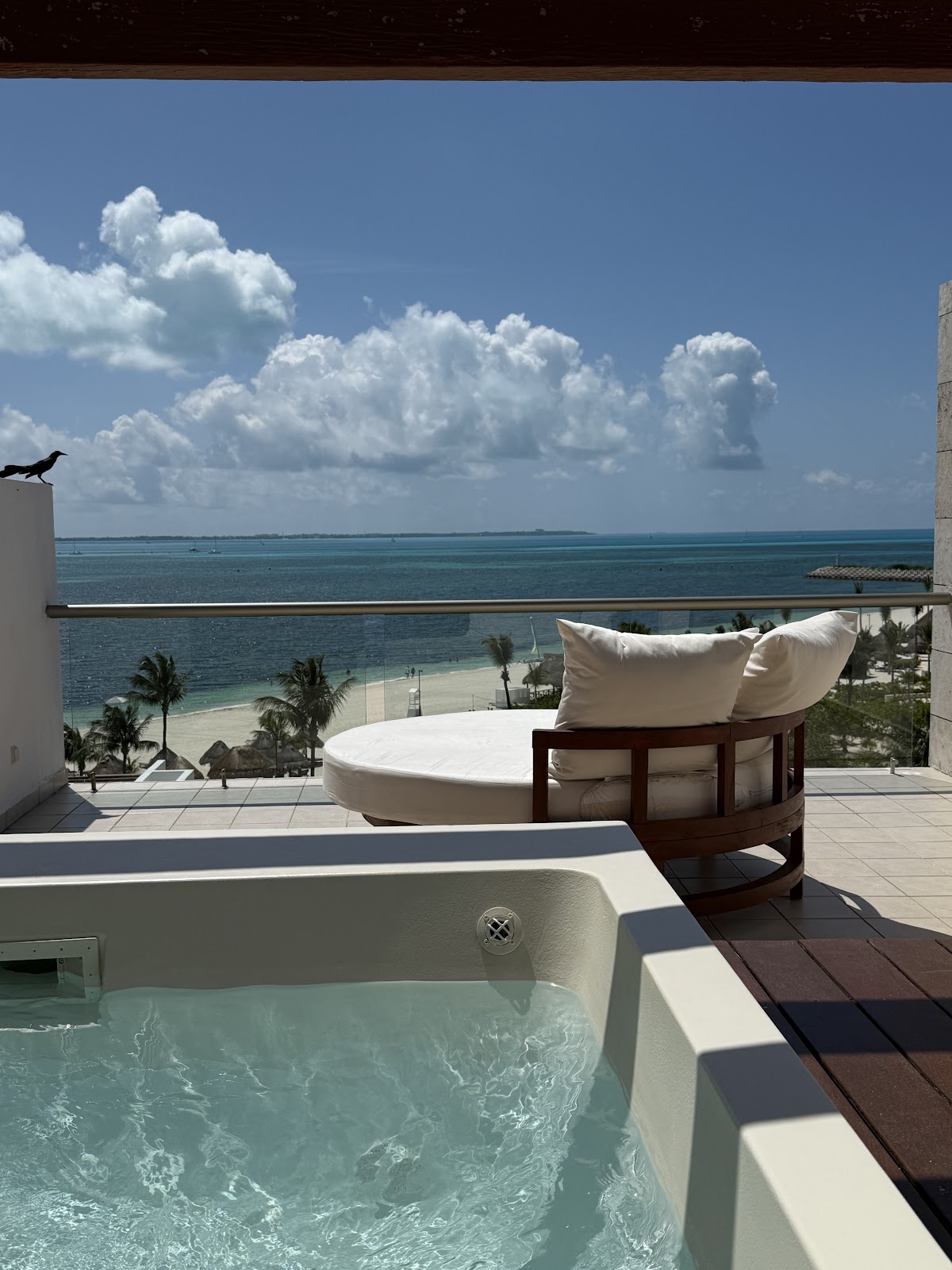 Hot tub and lounge bed on an oceanfront terrace at Excellence Playa Mujeres, with turquoise water and palm trees below.