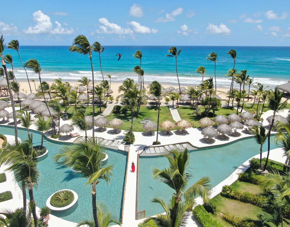Excellence Punta Cana pool and beach with turquoise water, palm trees, thatched umbrellas, and ocean beyond.