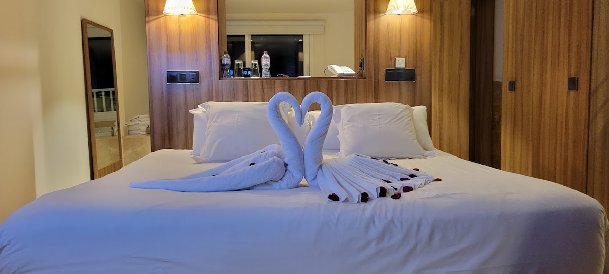 Two blue towels folded into swans with hearts facing each other on a bed in an Excellence Punta Cana guestroom with wood accent wall and mirror.