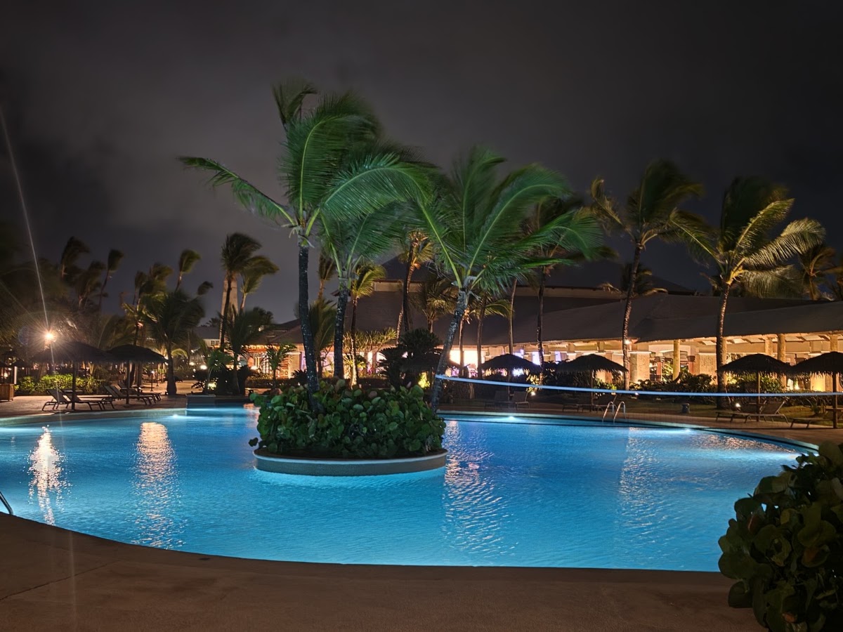 Illuminated pool with palm trees and landscaping at night, lit resort buildings and umbrellas visible in background.