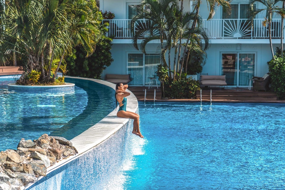 Guest sits on pool edge at Excellence Punta Cana with turquoise water, palm trees, and light blue building with balconies in background.