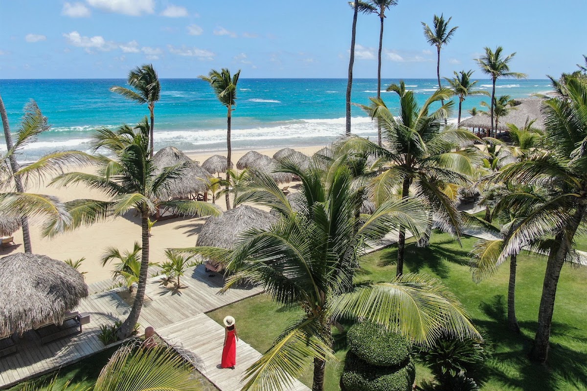 Woman in red dress stands on wooden boardwalk among palm trees overlooking white sand beach and turquoise ocean at Excellence Punta Cana.