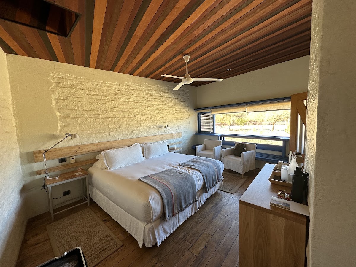 Resort bedroom with wooden ceiling, white textured accent wall, queen bed with white linens, two armchairs, and large window overlooking grounds.