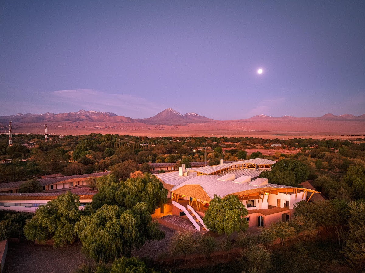 Resort buildings and pools at dusk with snow-capped volcanoes and the moon visible across the desert landscape of northern Chile.